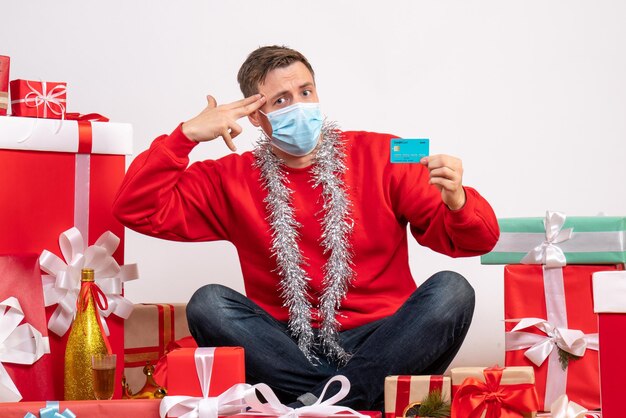 Vooraanzicht van een jonge man met een masker die rond kerstcadeautjes zit met een bankkaart op een witte muur