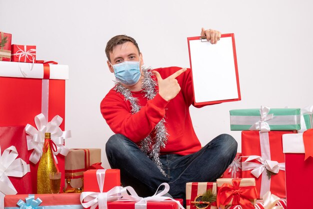 Gratis foto vooraanzicht van een jonge man met een masker die rond kerstcadeautjes zit met een opmerking op een witte muur