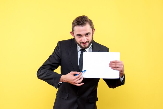 Vooraanzicht van een jonge man zakenman in een pak wijzend met een pen naar een blanco papier op geel