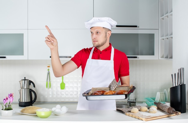 Vooraanzicht van een jonge mannelijke chef-kok die een houder draagt die versgebakken gebakjes vasthoudt en omhoog wijst in de witte keuken