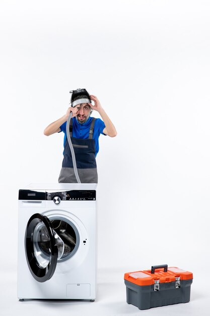 Vooraanzicht van een jonge reparateur in uniform die achter een wasmachine staat die de zak met pijpgereedschap op de muur blaast. op witte muur