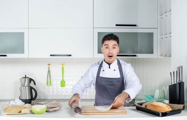 Vooraanzicht van een jonge verraste commischef in uniform die houder draagt en gebak bereidt in de witte keuken