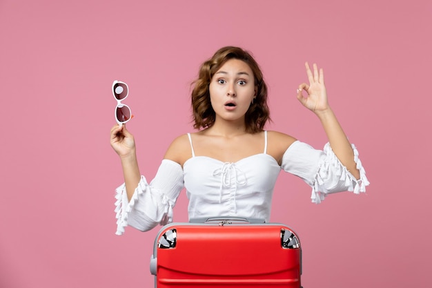 Vooraanzicht van een jonge vrouw die zich voordeed en zich voorbereidt op een reis met een rode tas op de roze muur