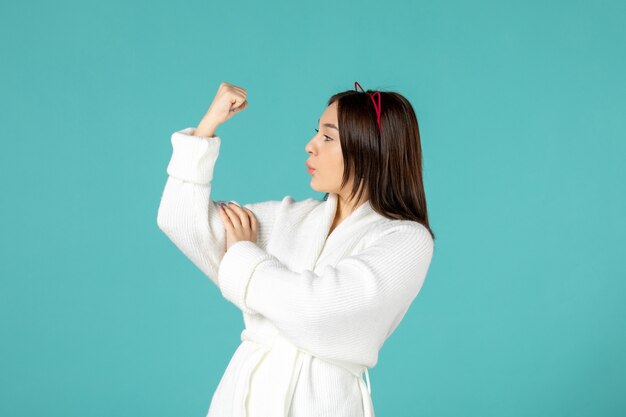 vooraanzicht van een jonge vrouw in een badjas die haar sterke arm op een blauwe muur laat zien