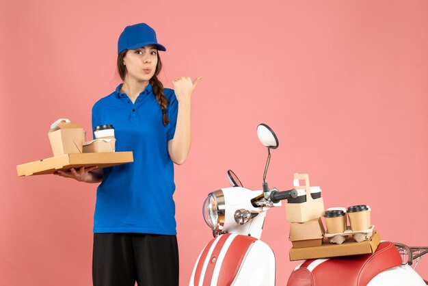 Vooraanzicht van een koeriersdame die naast een motorfiets staat met koffie en kleine cakes die terugwijst op een pastelkleurige perzikkleurige achtergrond