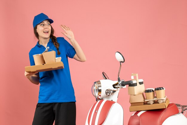 Vooraanzicht van een koeriersdame die naast een motorfiets staat met koffie en kleine taarten op een pastelkleurige perzikkleurige achtergrond