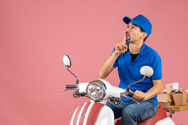 Vooraanzicht van een koeriersman met een hoed die op een scooter zit en een stiltegebaar maakt op een pastelkleurige perzikachtergrond