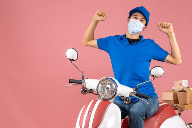 Vooraanzicht van een koeriersman met een medisch masker met een hoed die op een scooter zit en fronst op een pastelkleurige perzikachtergrond