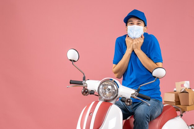 Vooraanzicht van een koeriersman met een medisch masker met een hoed die op een scooter zit en naar iets kijkt met een lachende gezichtsuitdrukking op een pastelkleurige perzikachtergrond