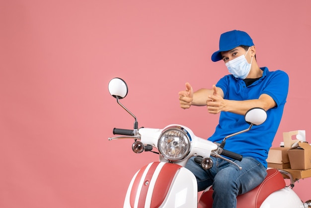 Vooraanzicht van een koeriersman met een medisch masker met een hoed die op een scooter zit en naar voren wijst op een pastelkleurige perzikachtergrond