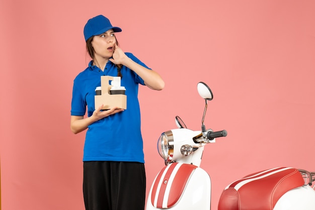 Vooraanzicht van een koeriersmeisje dat naast een motorfiets staat en koffie vasthoudt en diep nadenkt over een pastelkleurige perzikkleurige achtergrond