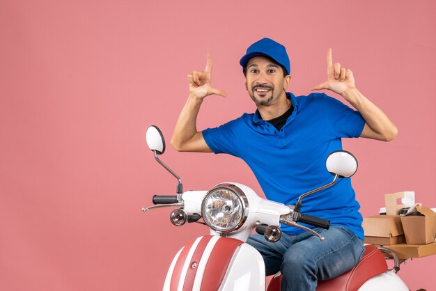 Vooraanzicht van een lachende en gelukkige koeriersman met een hoed op een scooter op een pastelkleurige perzikachtergrond peach