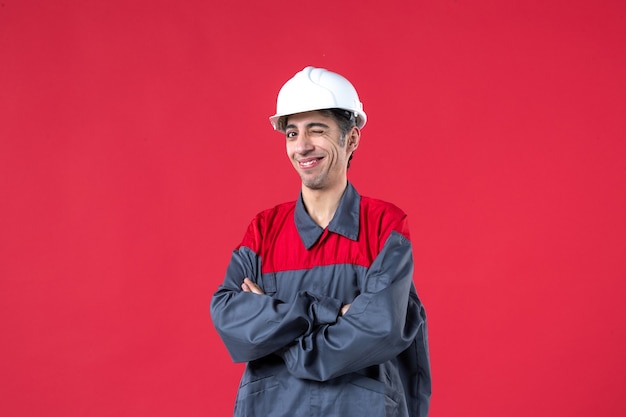 Vooraanzicht van een lachende jonge bouwer in uniform met een helm en zijn armen kruisend op een geïsoleerde rode muur