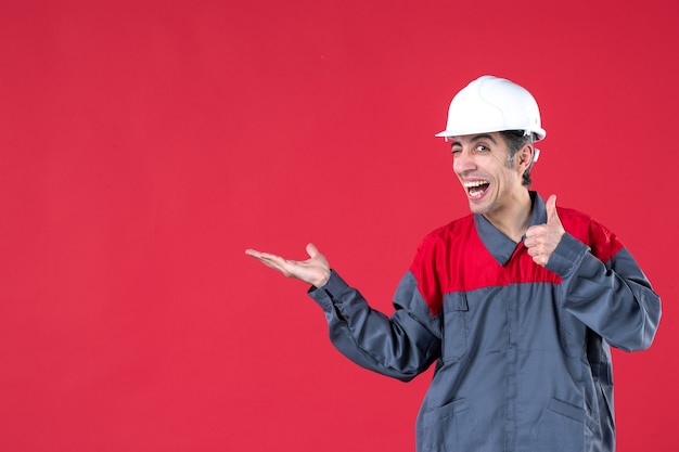 Vooraanzicht van een lachende jonge werknemer in uniform met een helm die iets wijst en een goed gebaar maakt op een geïsoleerde rode muur