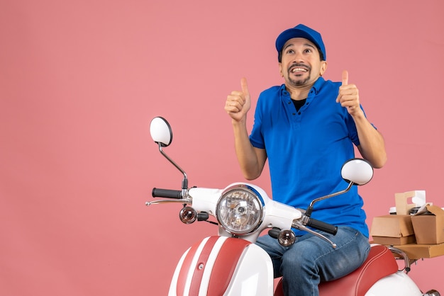 Vooraanzicht van een lachende koeriersman met een hoed die op een scooter zit en een goed gebaar maakt op een pastelkleurige perzikachtergrond