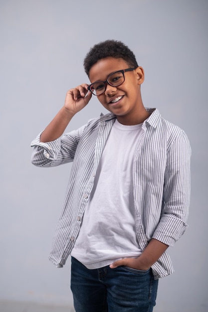 Vooraanzicht van een lachende, vrolijke, schattige jongen gekleed in een gestreept shirt die zijn bril met één hand aanraakt