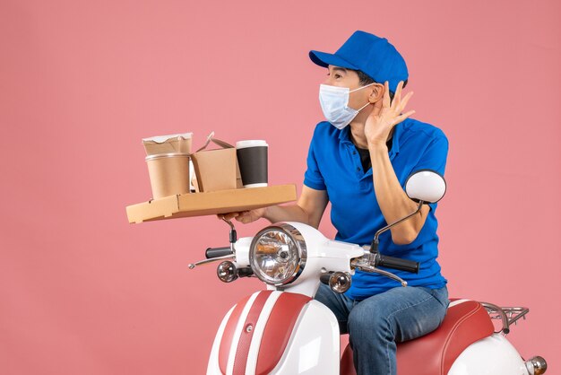 Vooraanzicht van een mannelijke bezorger met een masker met een hoed op een scooter die bestellingen aflevert en luistert naar de laatste roddels op een pastelkleurige perzikachtergrond