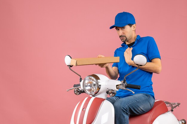 Vooraanzicht van een onzekere koeriersman met een hoed die op een scooter zit en de bestelling vasthoudt op een pastelkleurige perzikachtergrond