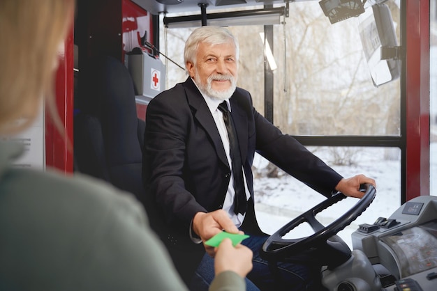 Vooraanzicht van een oudere lachende chauffeur in een zwarte jas met een ticket en een oogst van een vrouwelijke passagier