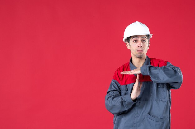 Vooraanzicht van een overstuur jonge architect in uniform met een helm die een stopgebaar maakt op een geïsoleerde rode muur