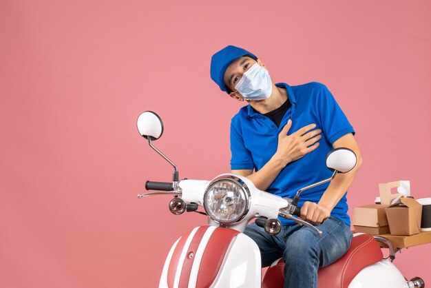 Vooraanzicht van een positieve koeriersman met een medisch masker met een hoed op een scooter op een pastelkleurige perzikachtergrond