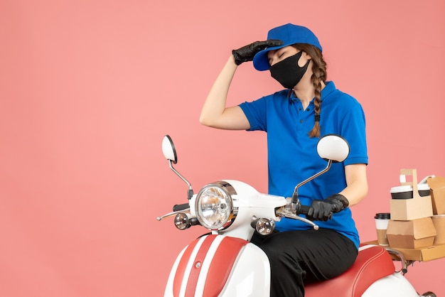 Vooraanzicht van een uitgeput koeriersmeisje met een medisch masker en handschoenen die op een scooter zitten en bestellingen afleveren op een pastelkleurige perzikachtergrond