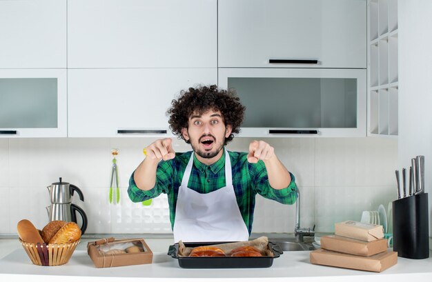 Vooraanzicht van een verbaasde man die achter een tafel staat met versgebakken gebak en naar voren wijst in de witte keuken