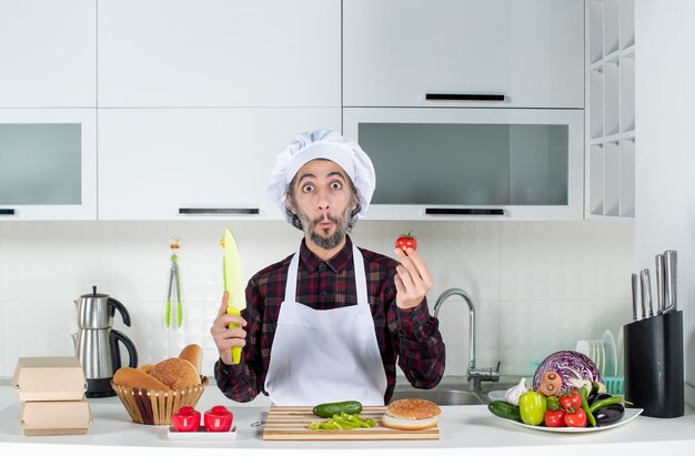 Vooraanzicht van een verbaasde mannelijke chef-kok met tomaat en mes in de keuken