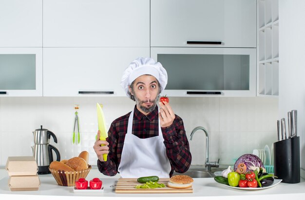Vooraanzicht van een verbaasde mannelijke chef-kok met tomaat en mes in de keuken
