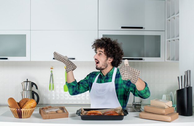 Vooraanzicht van een verraste jonge kerel met een houder die achter een tafel staat met vers gebakken gebak erop in de witte keuken