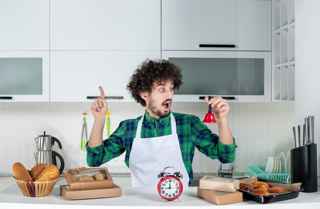 Vooraanzicht van een verraste jonge man die achter de tafel staat met verschillende gebakjes erop en met een rode bel die naar boven wijst in de witte keuken