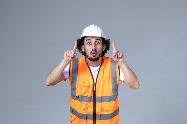 Vooraanzicht van een verraste mannelijke bouwer die een waarschuwingsvest met veiligheidshelm draagt en naar boven wijst op een grijze golfmuur