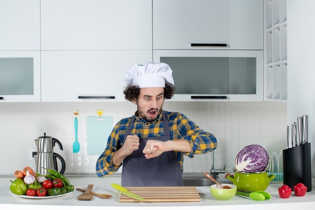 Vooraanzicht van een verraste mannelijke chef-kok met verse groenten en koken met keukengerei en zijn tijd in de witte keuken controleren