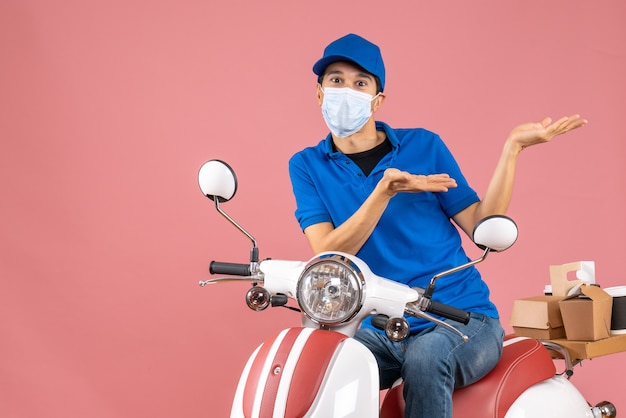 Vooraanzicht van een zich afvragende koeriersman met een medisch masker met een hoed op een scooter op een pastelkleurige perzikachtergrond