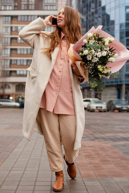 Vooraanzicht van elegante vrouw buitenshuis praten aan de telefoon en met een boeket bloemen