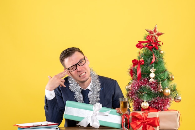 Vooraanzicht van gelukkig man vinger pistool zetten zijn tempel zittend aan de tafel in de buurt van kerstboom en presenteert op geel