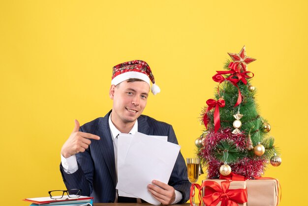 Vooraanzicht van gelukkig man wijzend op documenten zitten aan de tafel in de buurt van kerstboom en presenteert op geel