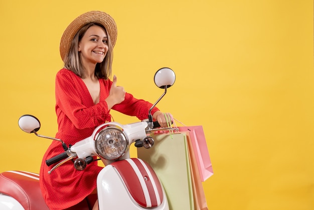 Vooraanzicht van gelukkige jonge vrouw in rode jurk op bromfiets bedrijf boodschappentassen duimen opgevend