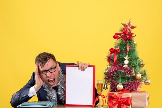 Vooraanzicht van geschreeuwde zakenman met Klembord zittend aan de tafel in de buurt van de kerstboom en presenteert op geel.