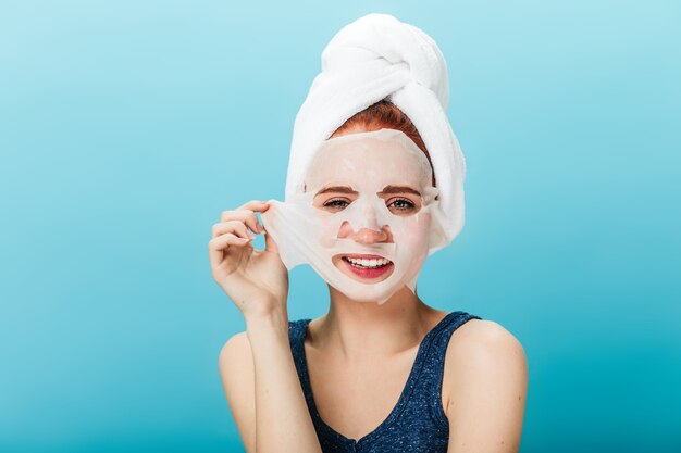 Vooraanzicht van glimlachend meisje gezichtsmasker opstijgen. Studio shot van zalige vrouw met handdoek op hoofd poseren op blauwe achtergrond.