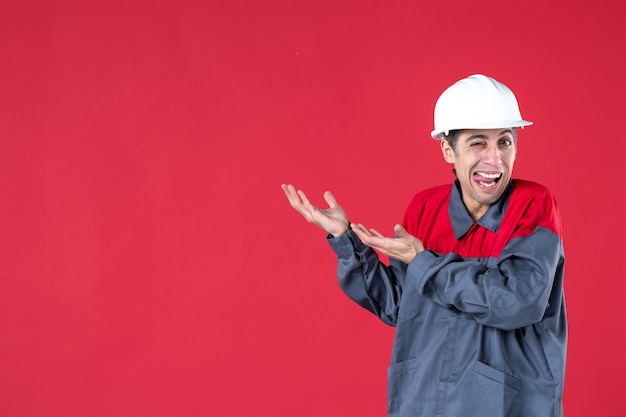 Vooraanzicht van grappige jonge werknemer in uniform met harde hoed en iets aan de rechterkant op geïsoleerde rode muur tonen