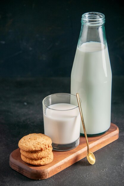 Vooraanzicht van heerlijke melk in glas en fles gouden lepel gestapelde koekjes op houten dienblad op donkere ondergrond