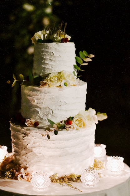 Vooraanzicht van heerlijke romige bruidstaart versierd met eucalyptus en witte rozen op tafel in de avond