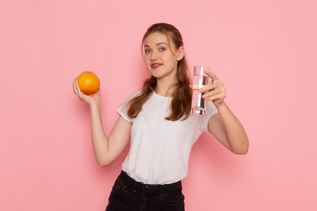 Vooraanzicht van jong wijfje dat in wit t-shirt verse grapefruit en glas water op de roze muur houdt