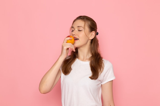 Vooraanzicht van jong wijfje dat in wit t-shirt verse perzik houdt die het op lichtroze muur eten