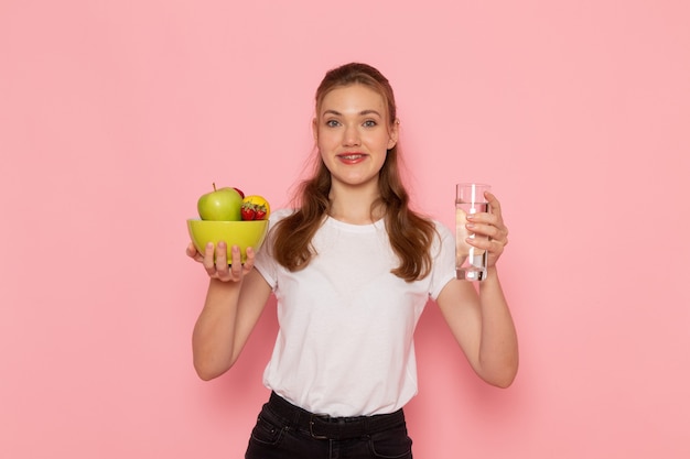 Vooraanzicht van jong wijfje in de witte plaat van de t-shirtholding met fruit en glas water op roze muur