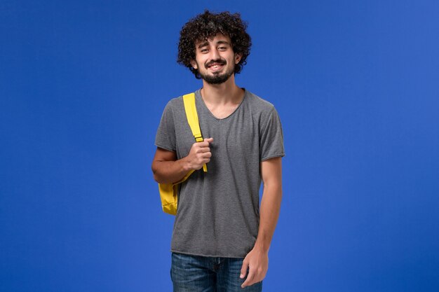 Vooraanzicht van jonge man in grijs t-shirt met gele rugzak lachend op de blauwe muur