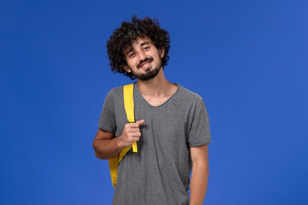 Vooraanzicht van jonge man in grijs t-shirt met gele rugzak lachend op de blauwe muur