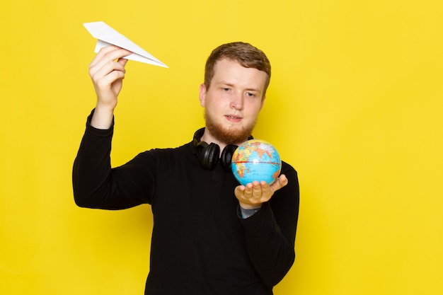 Vooraanzicht van jonge man in zwart shirt met papieren vliegtuigje en kleine wereldbol