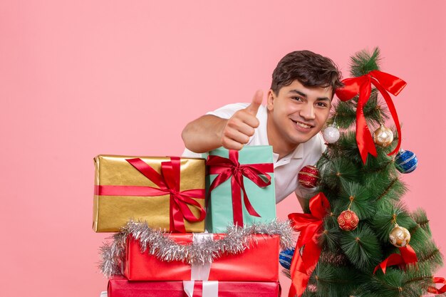 Vooraanzicht van jonge man rond kerstcadeautjes en vakantieboom op roze muur